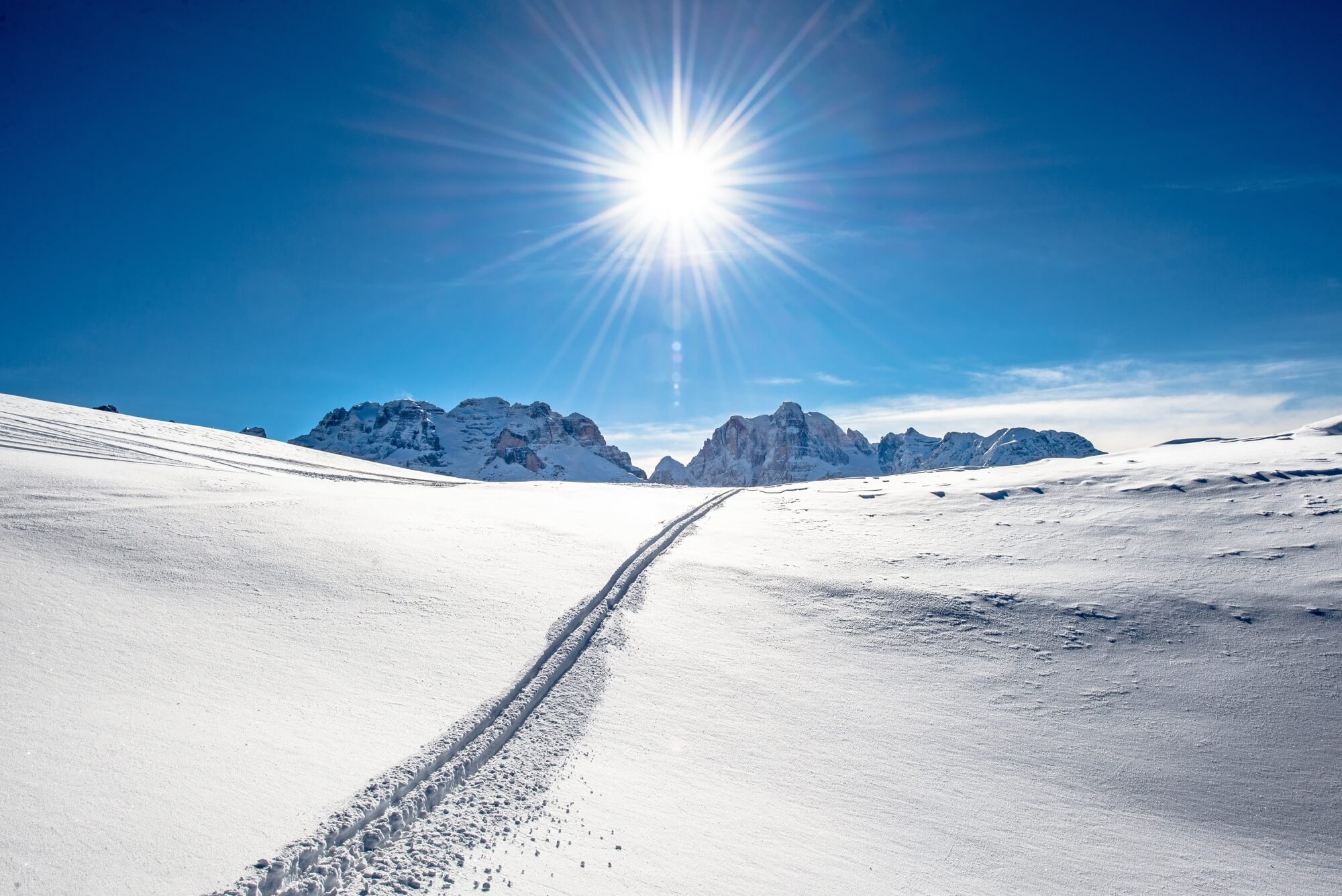 Paesaggio montano innevato con piste da sci e sole splendente.